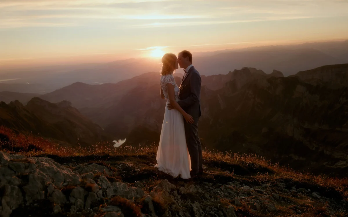 A bridal couple kisses at sunrise on Säntis, with the sun and Alpstein visible in the background.