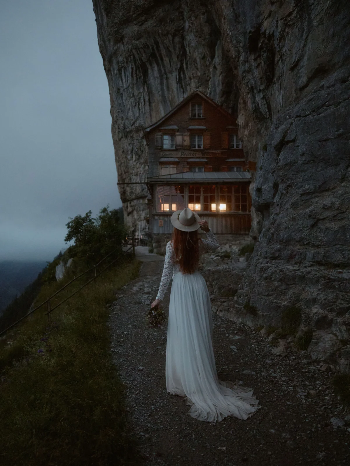 Braut mit Hut steht am Abend vor dem Berggasthaus Äscher, mystisches Licht.