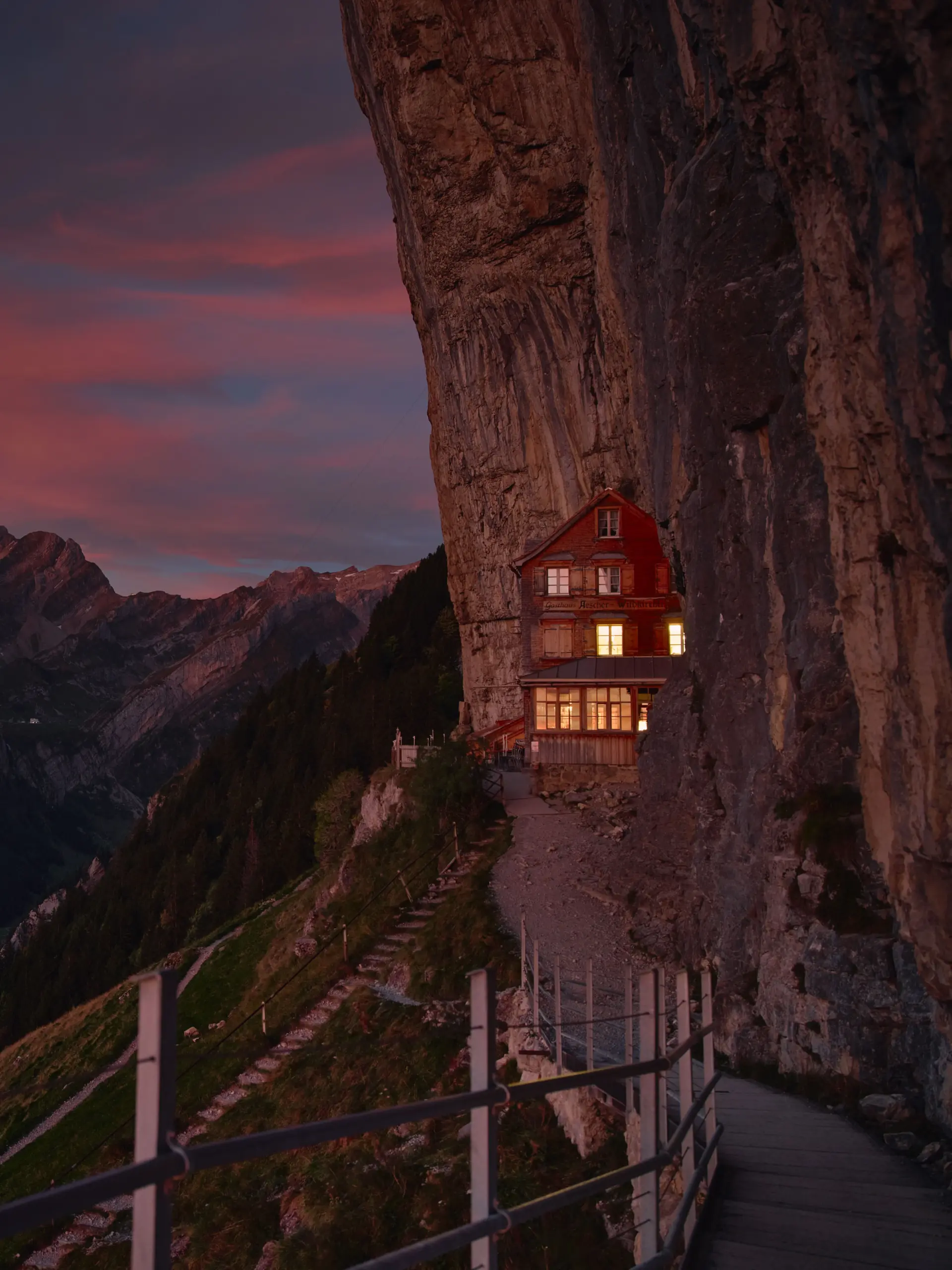 Das Berggasthaus Äscher bei Sonnenaufgang im Frühling, ein Gasthaus an einer Felswand. Der Himmel ist rosa.