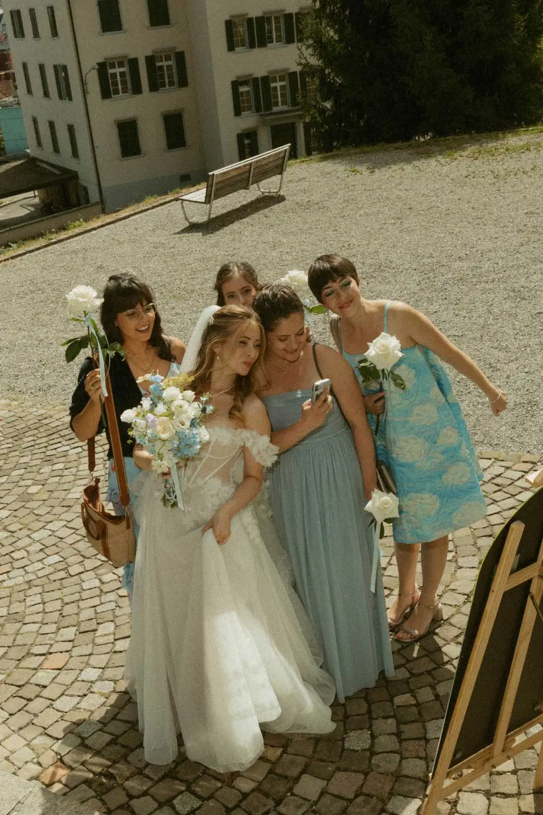 Braut lacht mit vier Freundinnen in blauen Kleidern auf dem Kopfsteinpflaster vor der Trauung