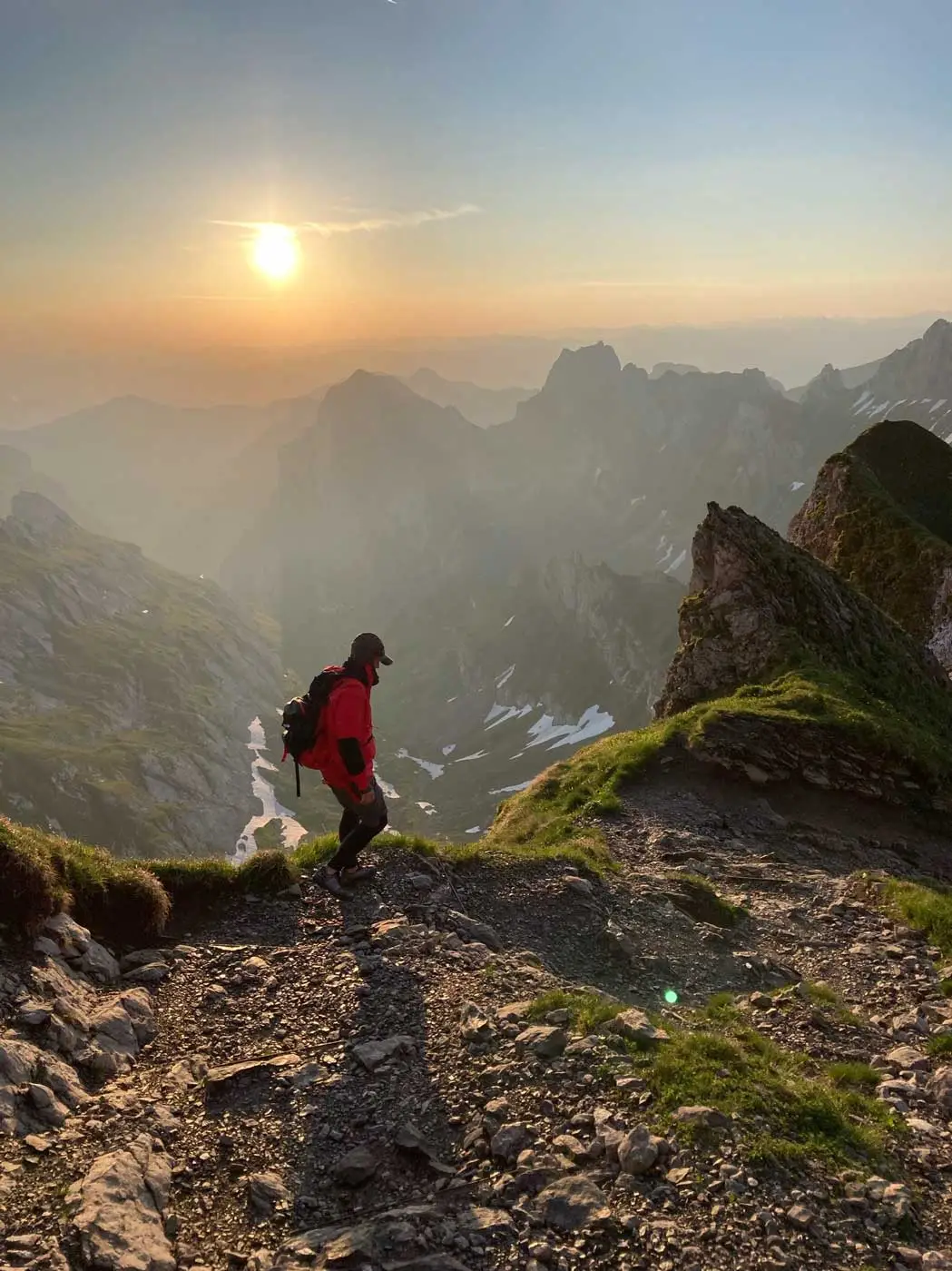 Hiker in a red jacket on a rocky Alpstein ridge, the sun low over the peaks, Appenzell
