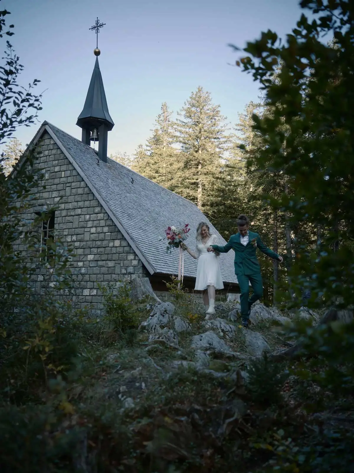 Brautpaar tritt mit Brautstrauss aus einer kleinen Holzschindel-Kapelle im Wald – Hochzeit in Appenzell