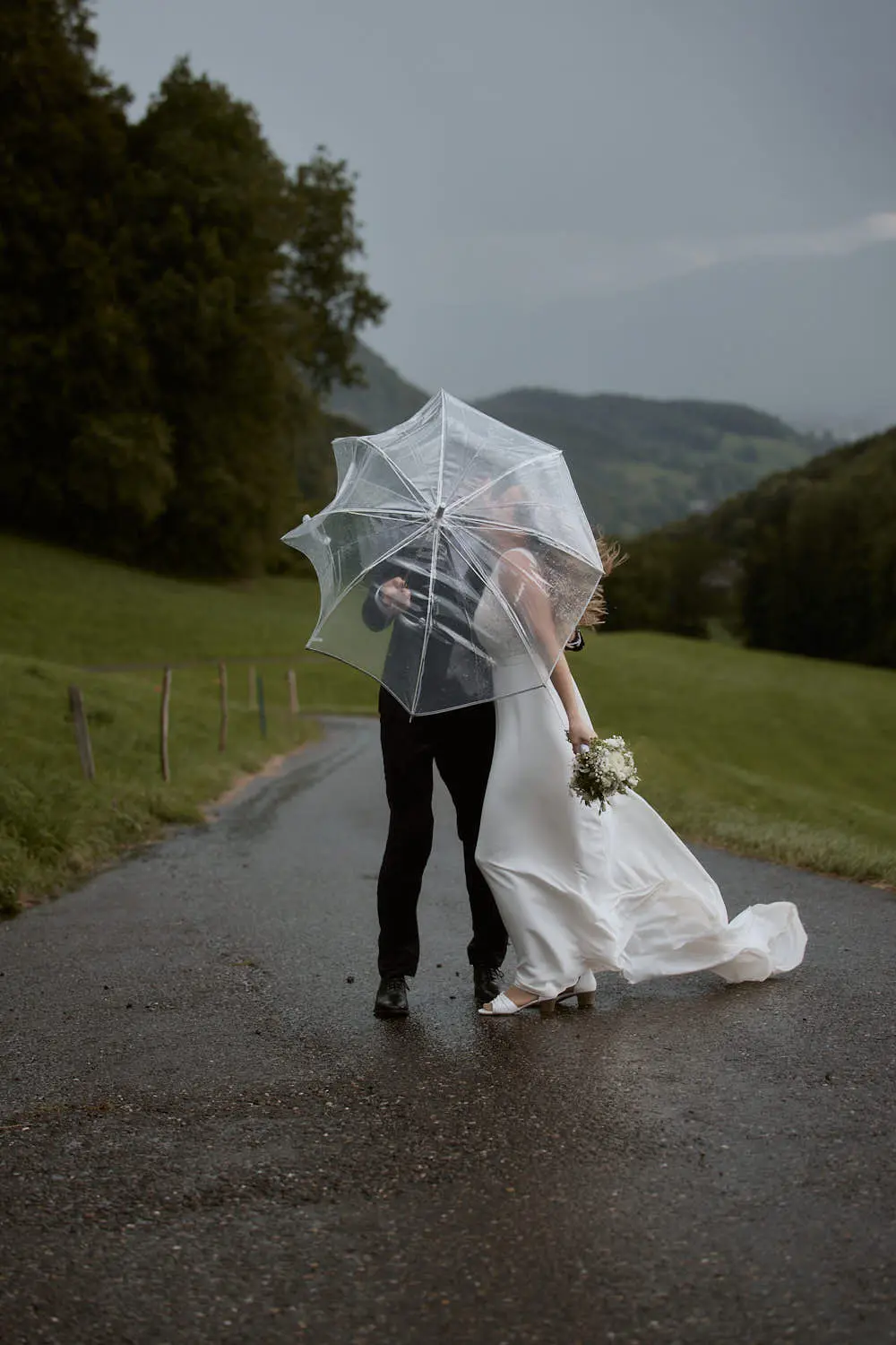 Brautpaar küsst sich unter einem Regenschirm auf einer nassen Strasse zwischen Wiesen in Malbun