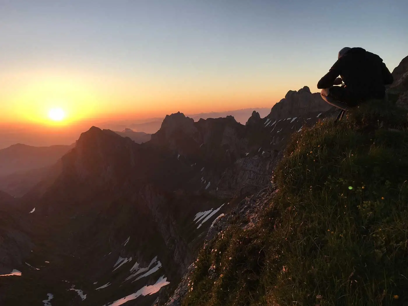 Photographer crouching on a grassy ridge above the Alpstein peaks, the sun low on the horizon